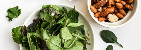 Top view of bowls containing plant-derived foods such as leafy greens, nuts, and spices on a white countertop