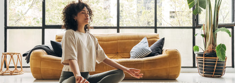 A woman with her eyes closed sits cross-legged on a rug and meditates in a sunny living room