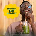 Person drinking green juice with a straw, wearing glasses, against a wooden background with 'Supports Gut Health' text.