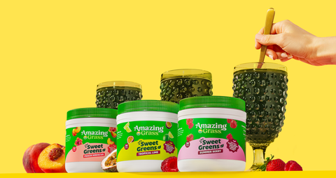 three jars of Amazing Grass Sweet Greens products next a glass of green juice with a hand stirring with a spoon and fruits scattered over a yellow background